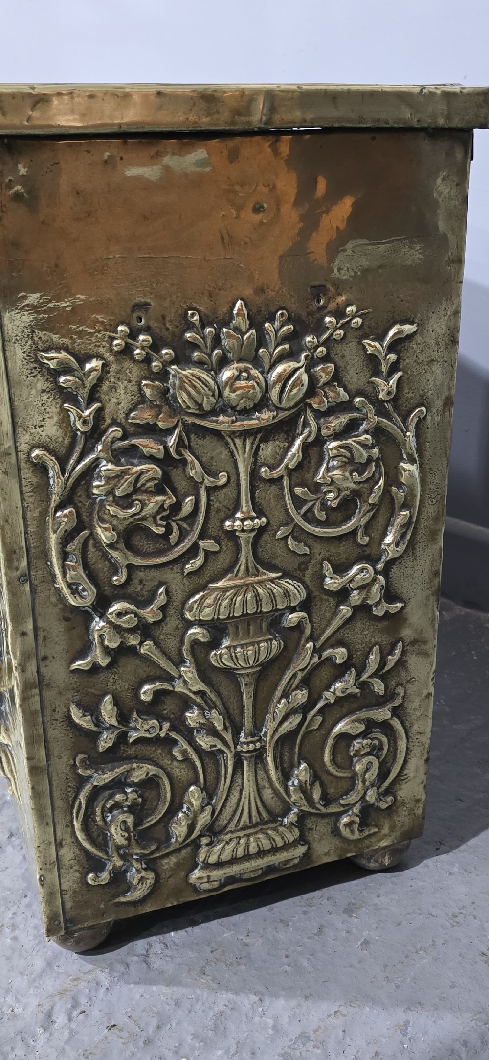 A rectangular Victorian brass log bin featuring detailed repousse relief panels of figures inside a domestic hearth interior. View VI