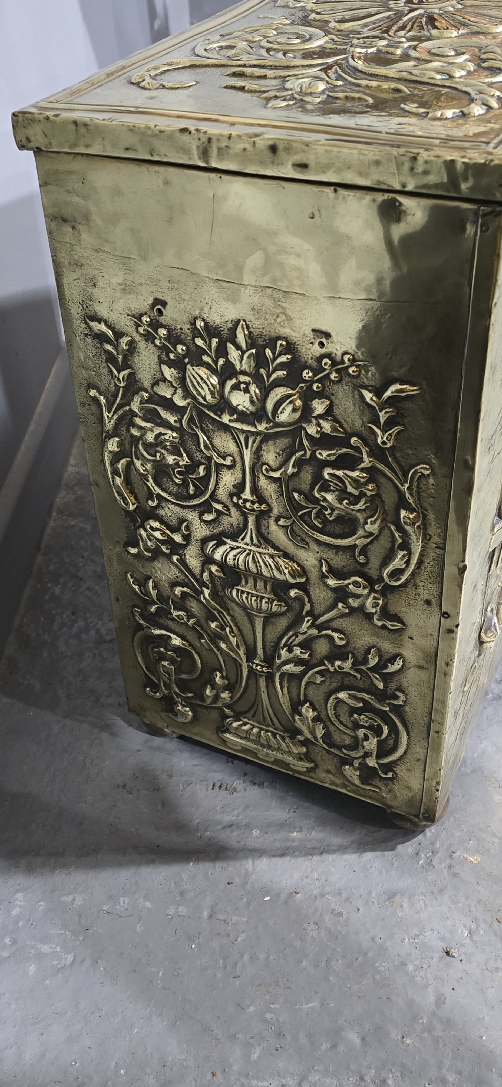 A rectangular Victorian brass log bin featuring detailed repousse relief panels of figures inside a domestic hearth interior. View VII
