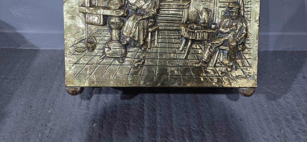 A rectangular Victorian brass log bin featuring detailed repousse relief panels of figures inside a domestic hearth interior. View X