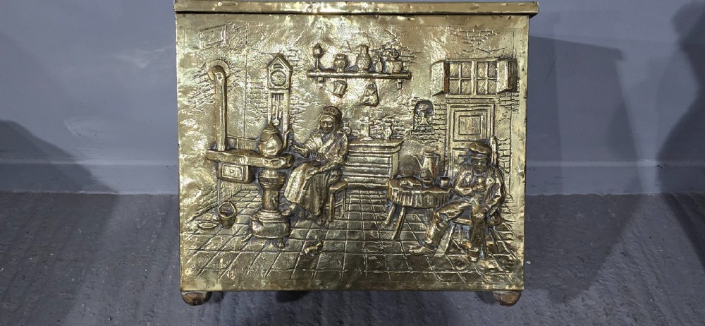 A rectangular Victorian brass log bin featuring detailed repousse relief panels of figures inside a domestic hearth interior. View XI
