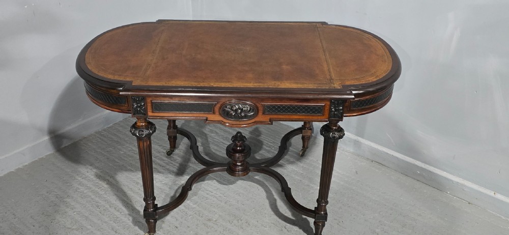 An antique dark brown wood library table featuring a leather inset top, carved frieze, turned legs, cross stretchers with a central finial, and brass castors. View II