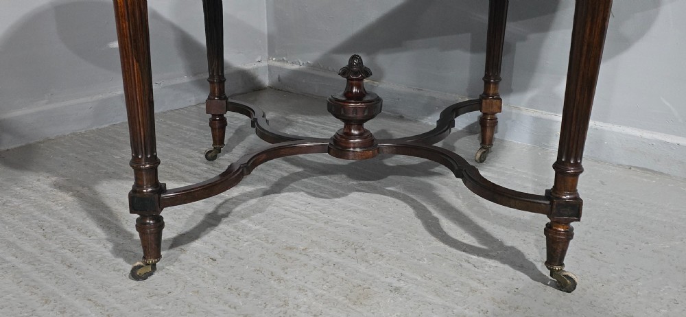 An antique dark brown wood library table featuring a leather inset top, carved frieze, turned legs, cross stretchers with a central finial, and brass castors. View III