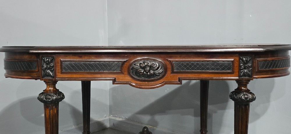An antique dark brown wood library table featuring a leather inset top, carved frieze, turned legs, cross stretchers with a central finial, and brass castors. View IV