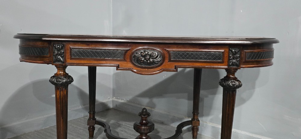 An antique dark brown wood library table featuring a leather inset top, carved frieze, turned legs, cross stretchers with a central finial, and brass castors. View V