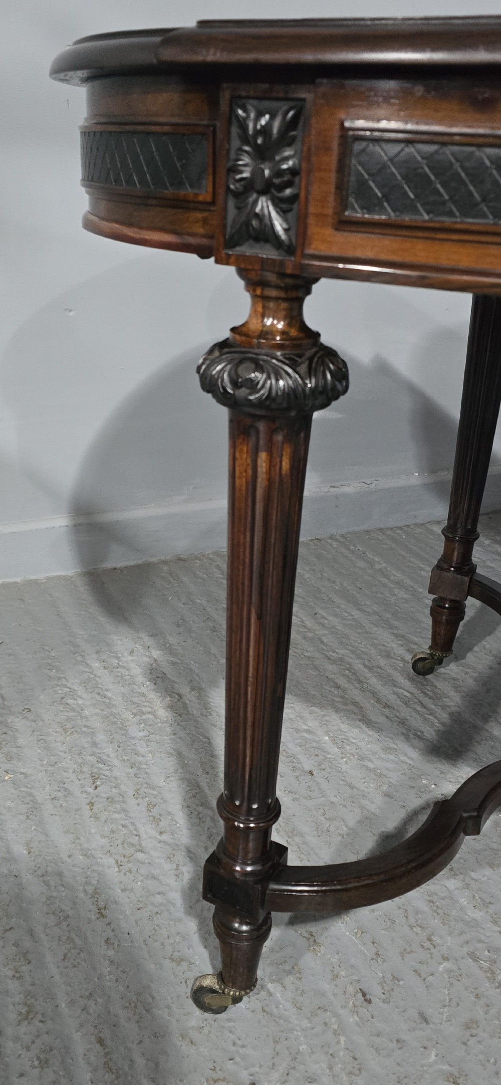 An antique dark brown wood library table featuring a leather inset top, carved frieze, turned legs, cross stretchers with a central finial, and brass castors. View VI