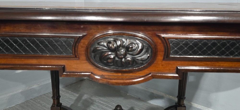 An antique dark brown wood library table featuring a leather inset top, carved frieze, turned legs, cross stretchers with a central finial, and brass castors. View VII