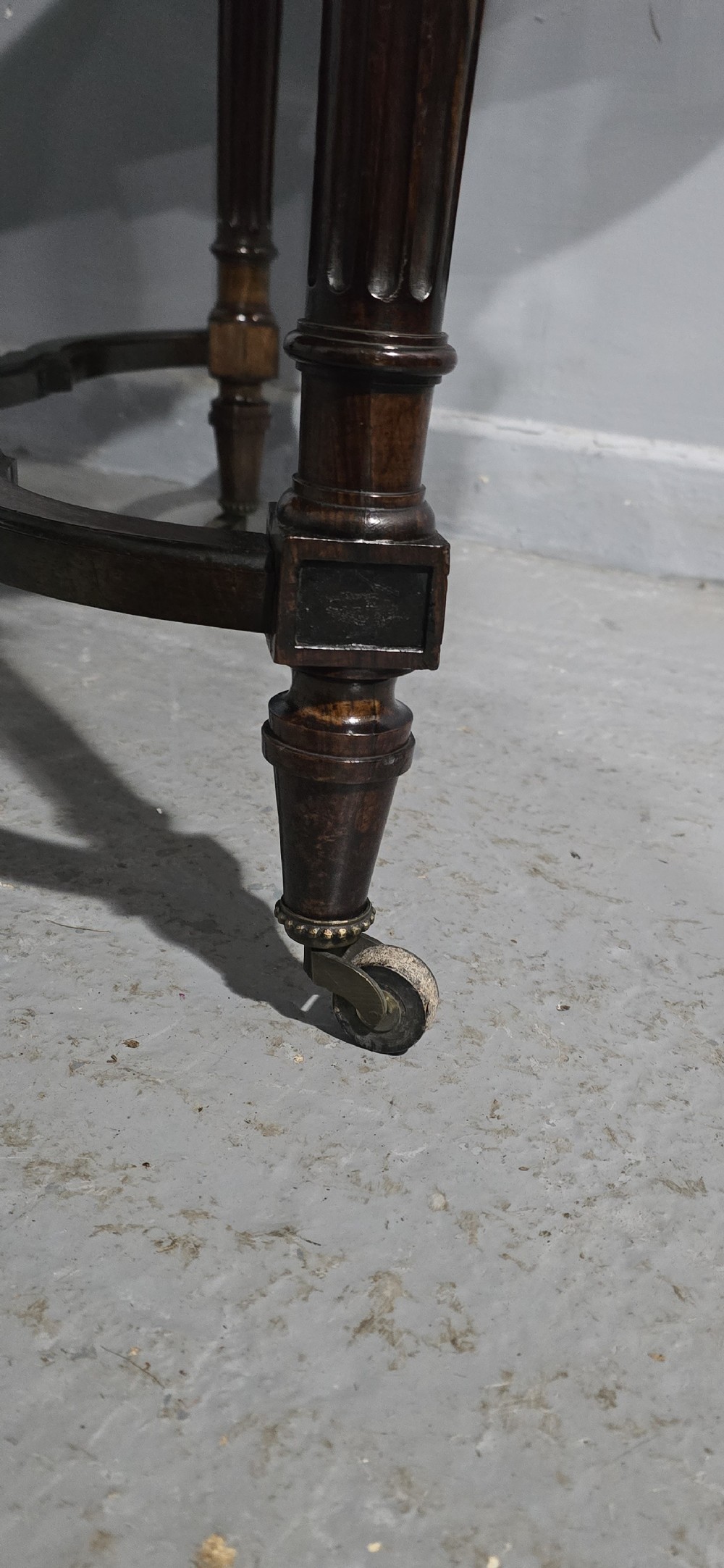 An antique dark brown wood library table featuring a leather inset top, carved frieze, turned legs, cross stretchers with a central finial, and brass castors. View IX