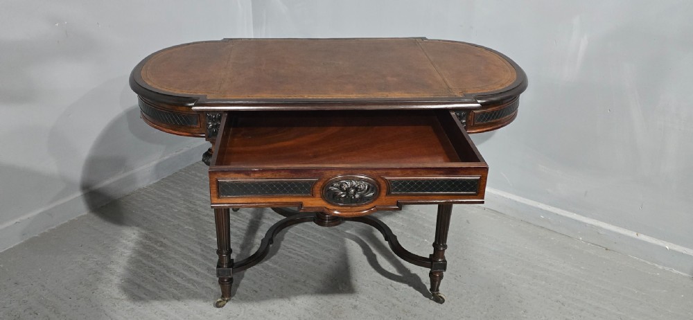 An antique dark brown wood library table featuring a leather inset top, carved frieze, turned legs, cross stretchers with a central finial, and brass castors. View XIII