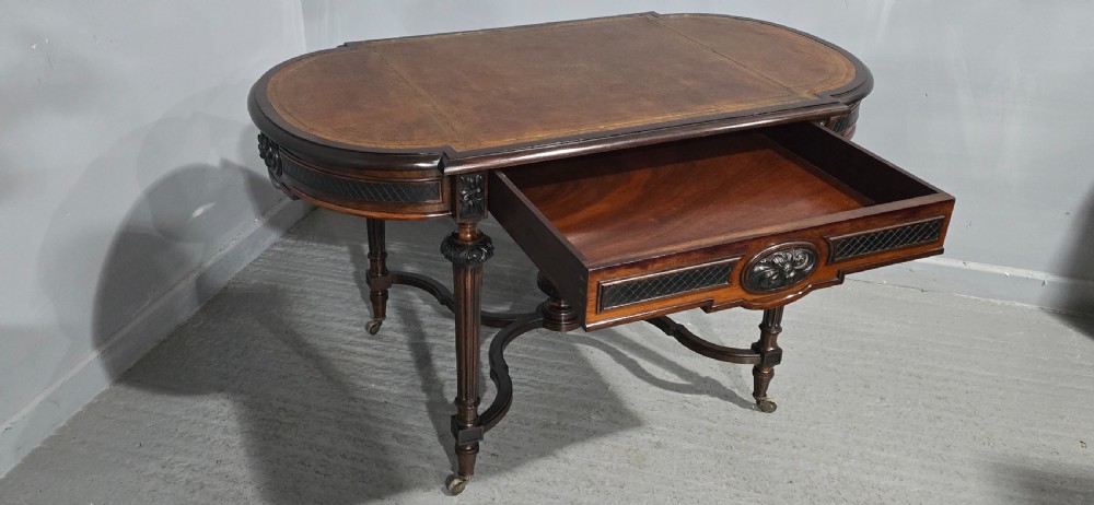 An antique dark brown wood library table featuring a leather inset top, carved frieze, turned legs, cross stretchers with a central finial, and brass castors. View XIV
