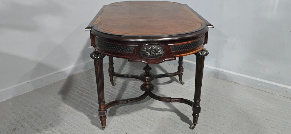 An antique dark brown wood library table featuring a leather inset top, carved frieze, turned legs, cross stretchers with a central finial, and brass castors. View XV