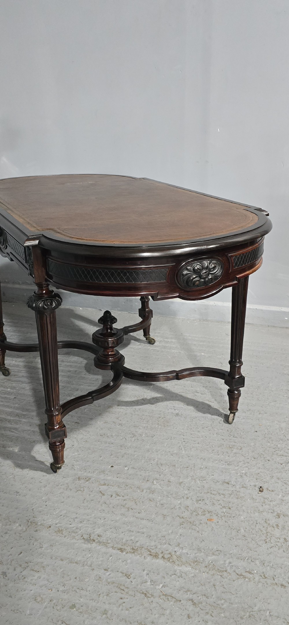 An antique dark brown wood library table featuring a leather inset top, carved frieze, turned legs, cross stretchers with a central finial, and brass castors. View XVI