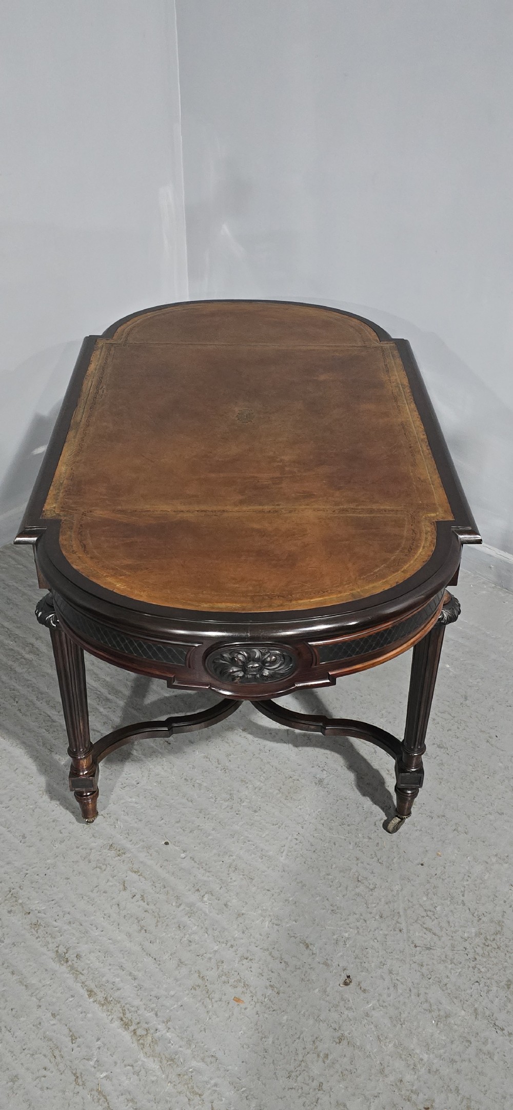 An antique dark brown wood library table featuring a leather inset top, carved frieze, turned legs, cross stretchers with a central finial, and brass castors. View XIX