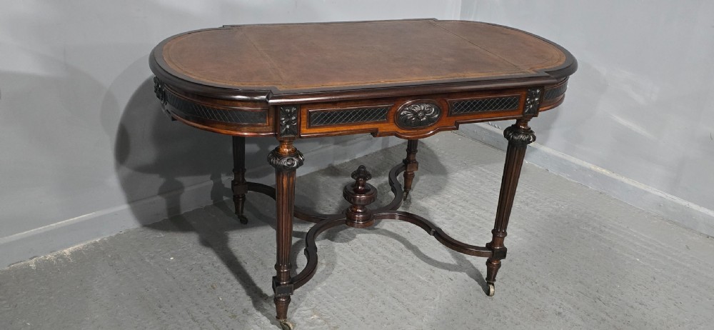 An antique dark brown wood library table featuring a leather inset top, carved frieze, turned legs, cross stretchers with a central finial, and brass castors. View XX