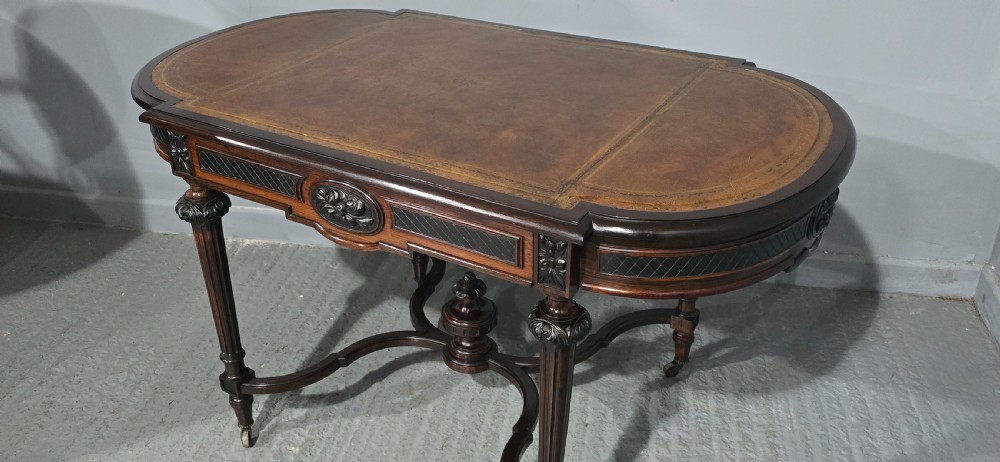 An antique dark brown wood library table featuring a leather inset top, carved frieze, turned legs, cross stretchers with a central finial, and brass castors. View XXI