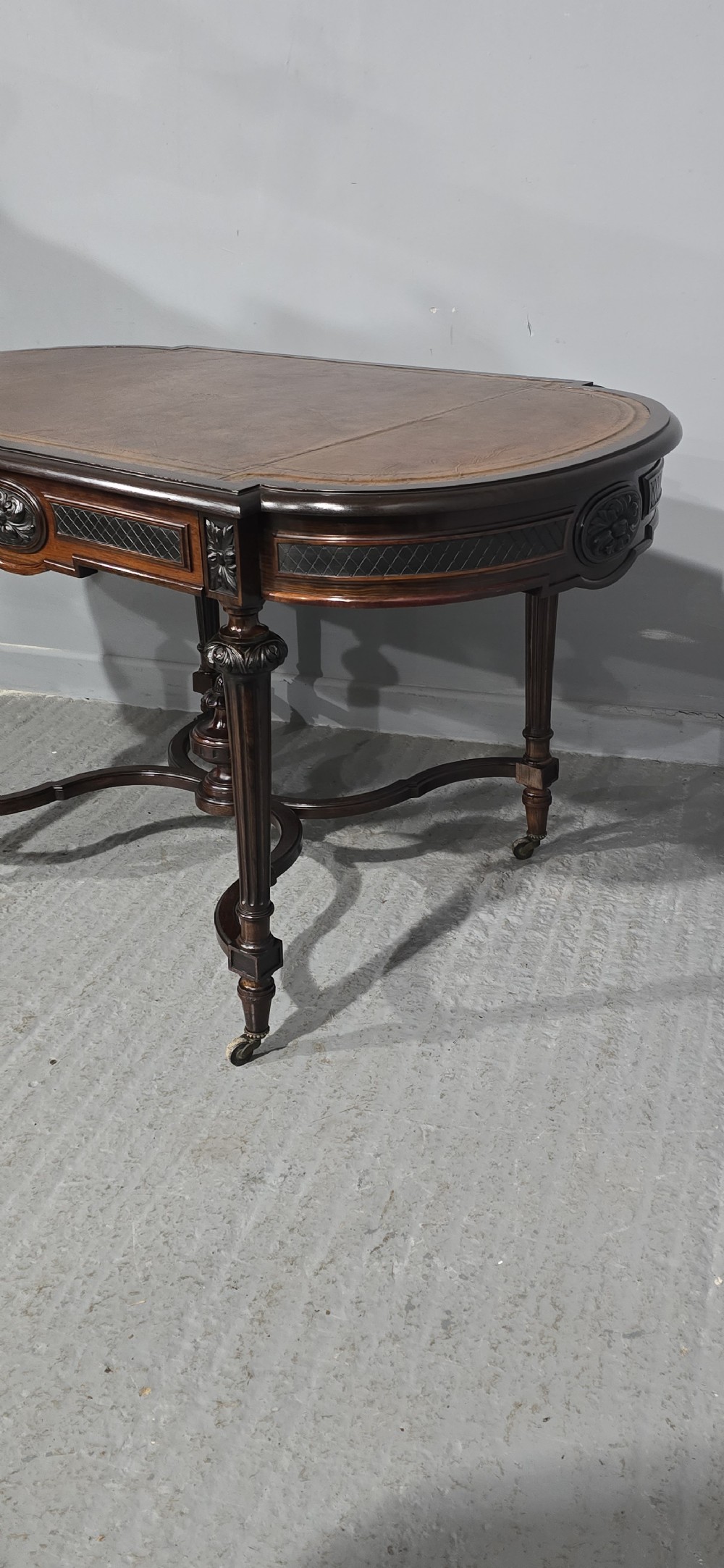 An antique dark brown wood library table featuring a leather inset top, carved frieze, turned legs, cross stretchers with a central finial, and brass castors. View XXII