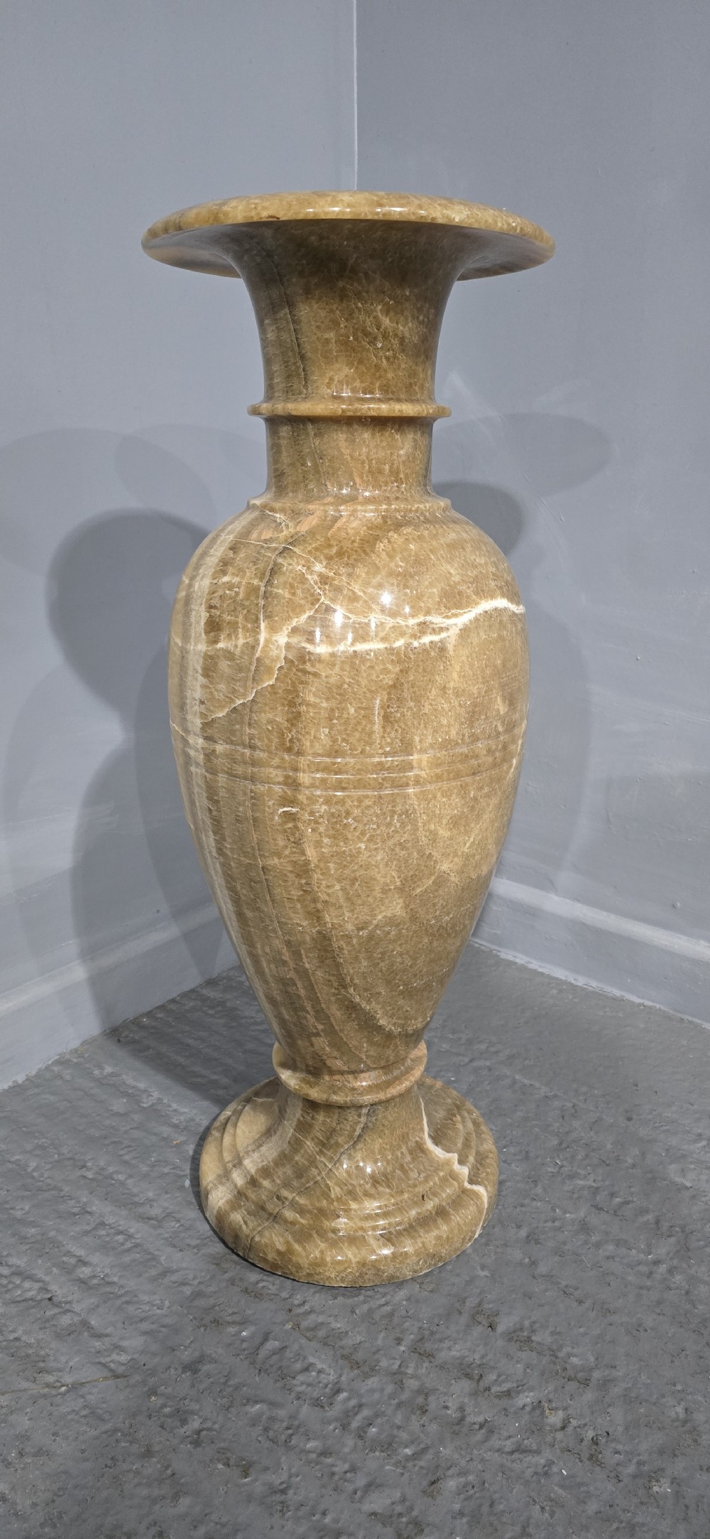 A large carved honey-brown alabaster vase featuring natural white veining, a flared rim, a knopped neck, and a stepped circular pedestal base. View III