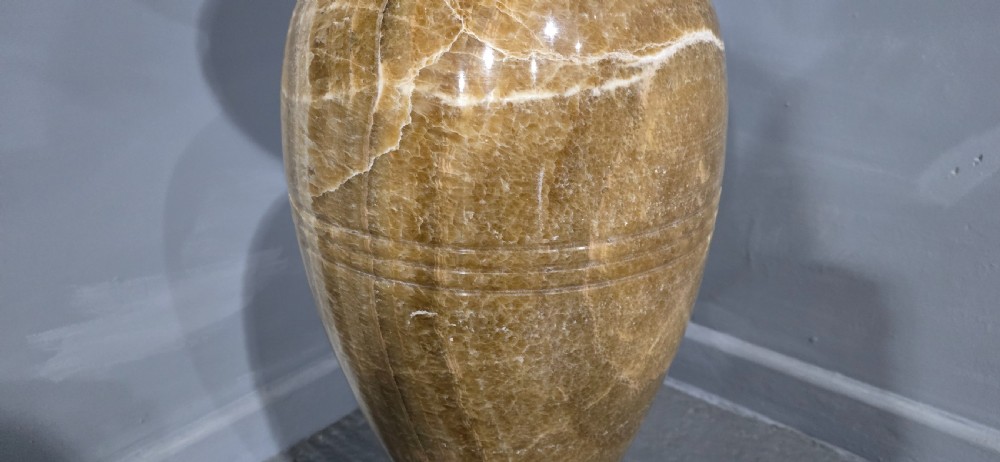 A large carved honey-brown alabaster vase featuring natural white veining, a flared rim, a knopped neck, and a stepped circular pedestal base. View IV