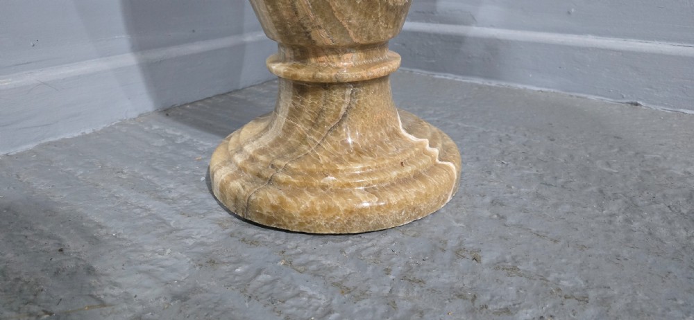 A large carved honey-brown alabaster vase featuring natural white veining, a flared rim, a knopped neck, and a stepped circular pedestal base. View V