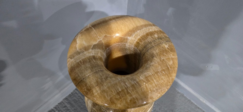 A large carved honey-brown alabaster vase featuring natural white veining, a flared rim, a knopped neck, and a stepped circular pedestal base. View VII