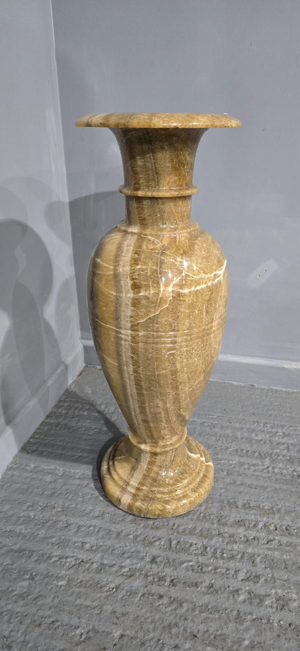 A large carved honey-brown alabaster vase featuring natural white veining, a flared rim, a knopped neck, and a stepped circular pedestal base. View IX