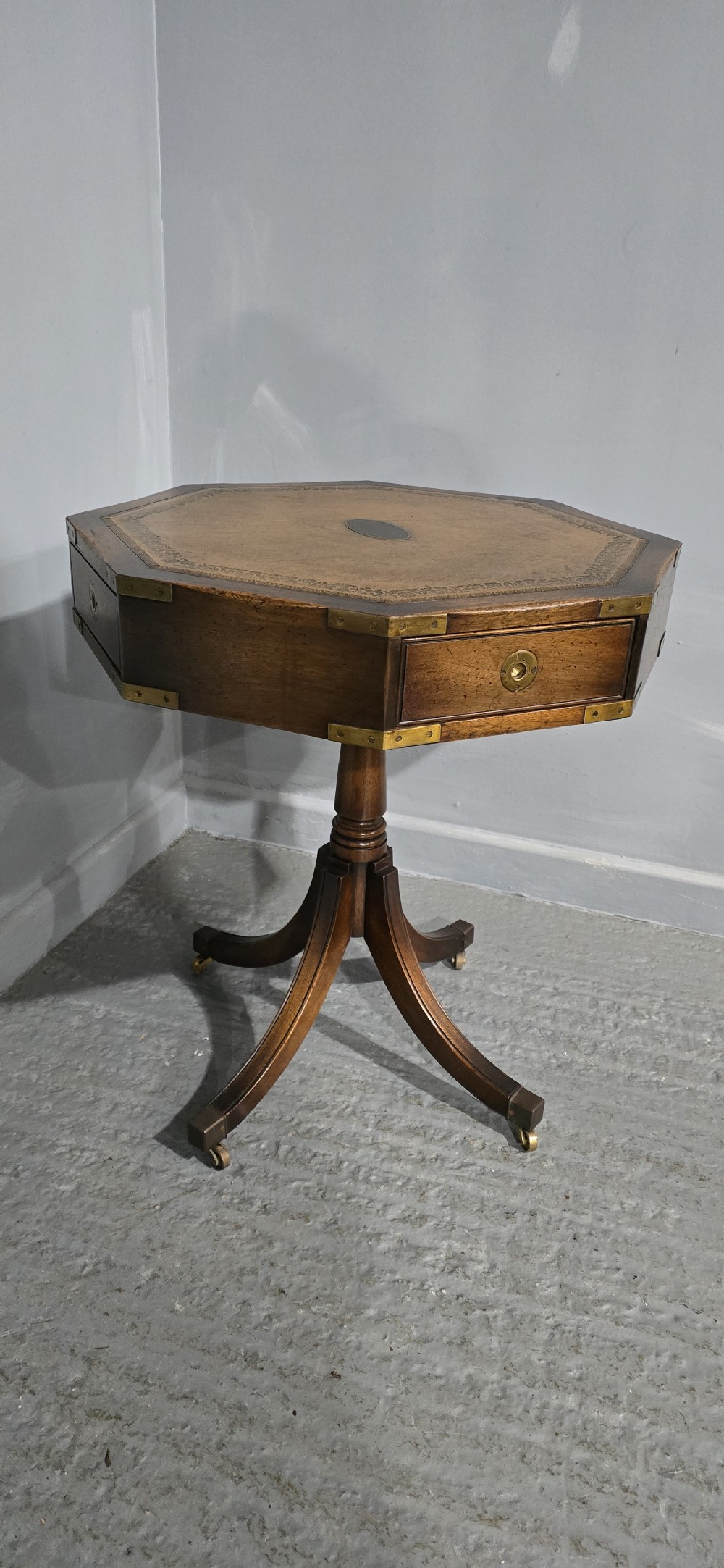 An octagonal mahogany campaign drum table with a gilt-tooled brown leather insert top, brass corner mounts, a single frieze drawer, and a four-legged pedestal base with brass castors. View II
