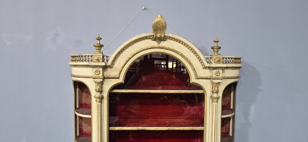 An antique French cream and gold painted wall vitrine featuring a central arched glazed door, open side shelves backed with red velvet, and carved gilt accents. View III