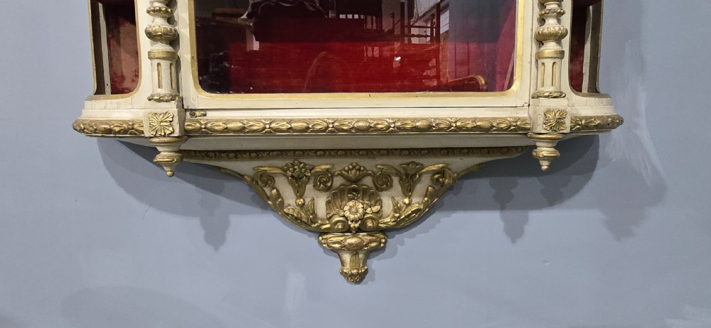 An antique French cream and gold painted wall vitrine featuring a central arched glazed door, open side shelves backed with red velvet, and carved gilt accents. View IV