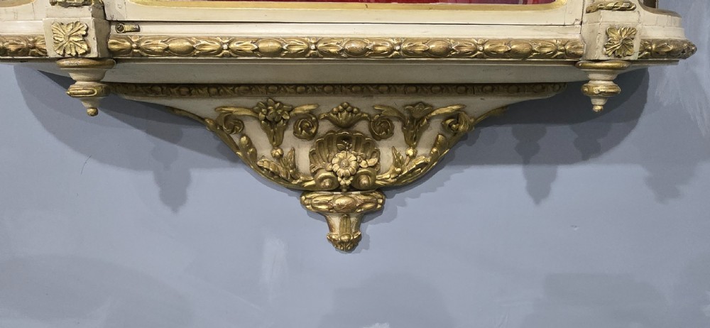 An antique French cream and gold painted wall vitrine featuring a central arched glazed door, open side shelves backed with red velvet, and carved gilt accents. View V