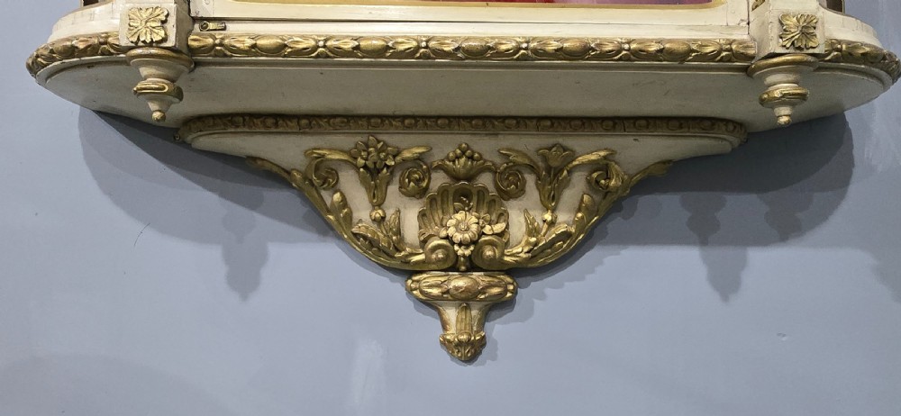 An antique French cream and gold painted wall vitrine featuring a central arched glazed door, open side shelves backed with red velvet, and carved gilt accents. View VI