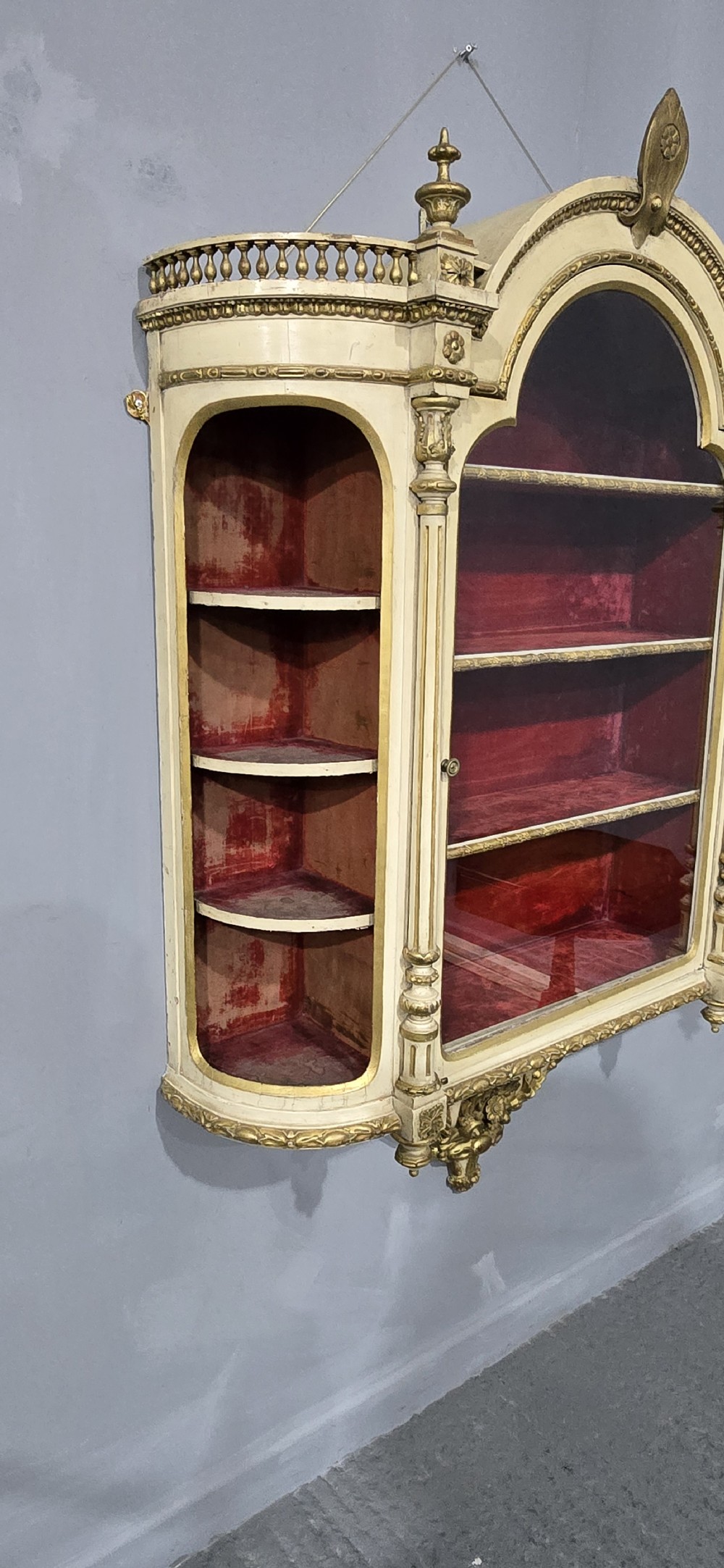 An antique French cream and gold painted wall vitrine featuring a central arched glazed door, open side shelves backed with red velvet, and carved gilt accents. View VIII