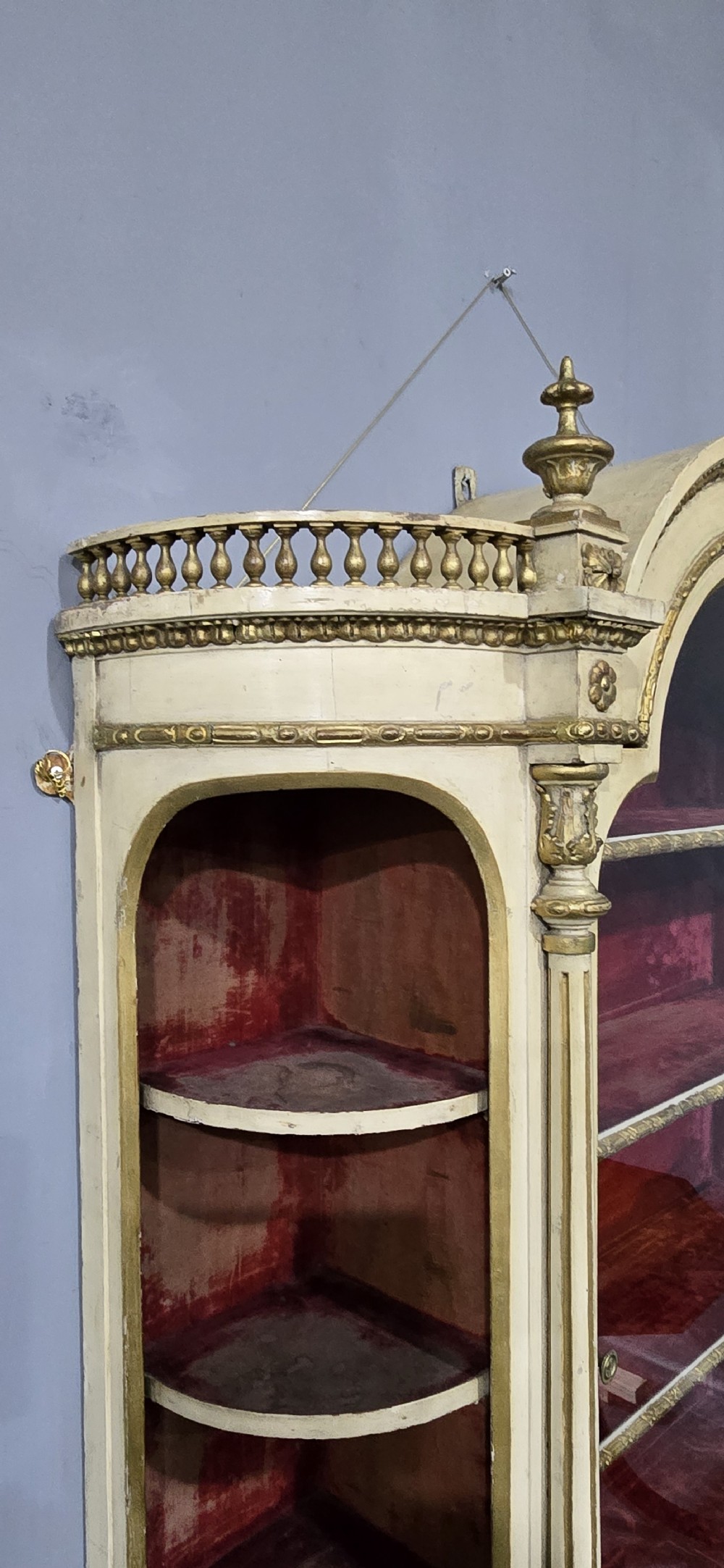 An antique French cream and gold painted wall vitrine featuring a central arched glazed door, open side shelves backed with red velvet, and carved gilt accents. View IX
