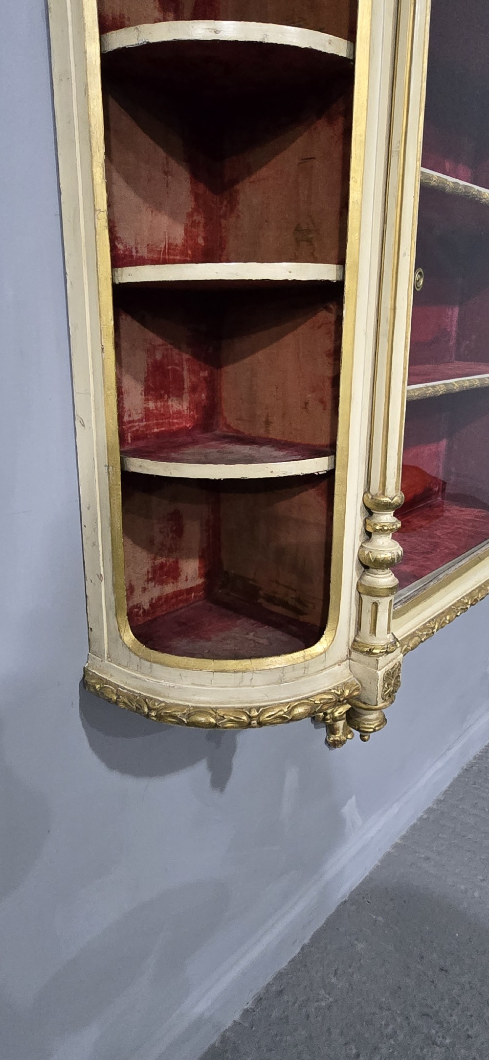 An antique French cream and gold painted wall vitrine featuring a central arched glazed door, open side shelves backed with red velvet, and carved gilt accents. View X