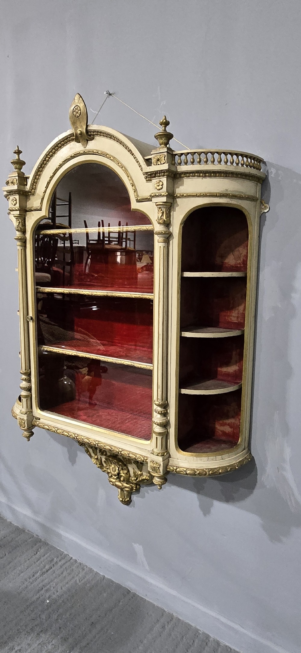An antique French cream and gold painted wall vitrine featuring a central arched glazed door, open side shelves backed with red velvet, and carved gilt accents. View XI