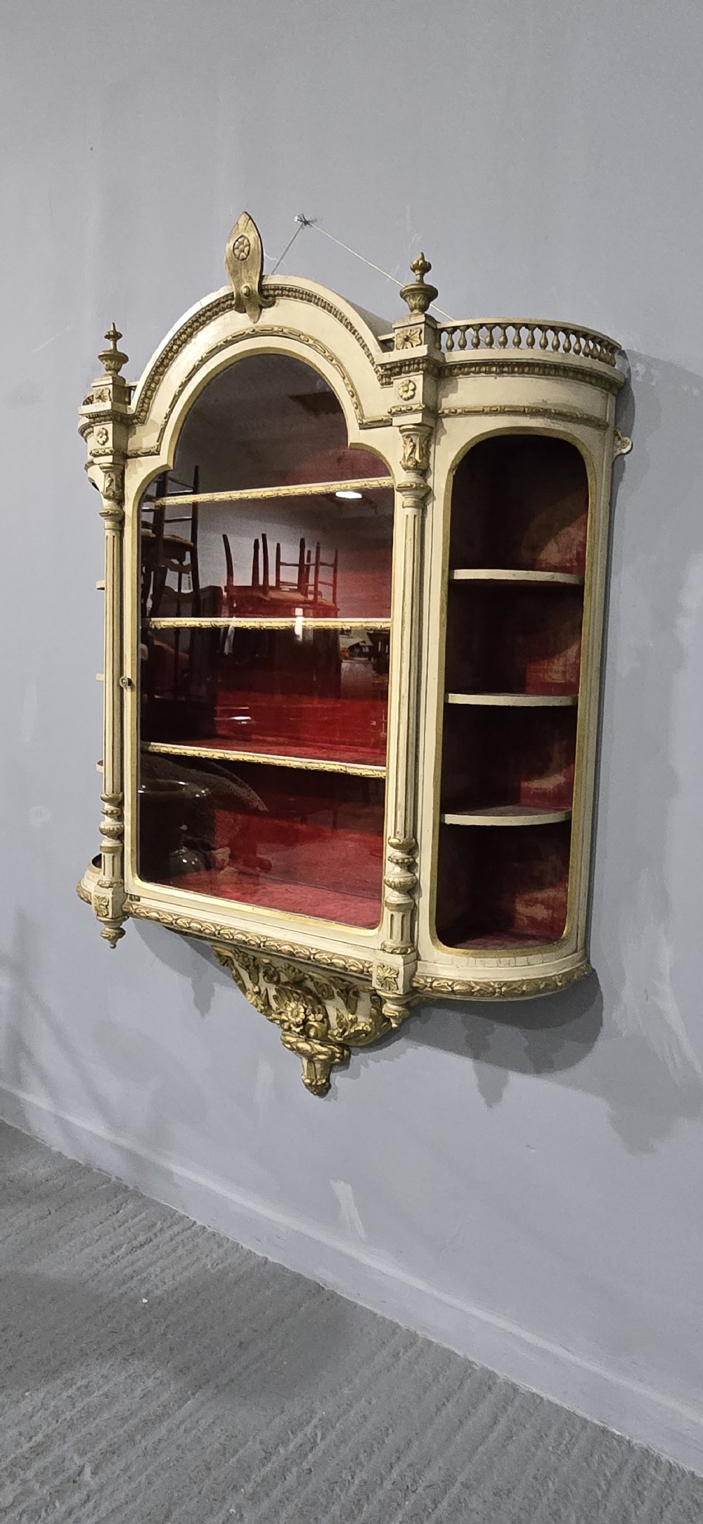 An antique French cream and gold painted wall vitrine featuring a central arched glazed door, open side shelves backed with red velvet, and carved gilt accents. View XII