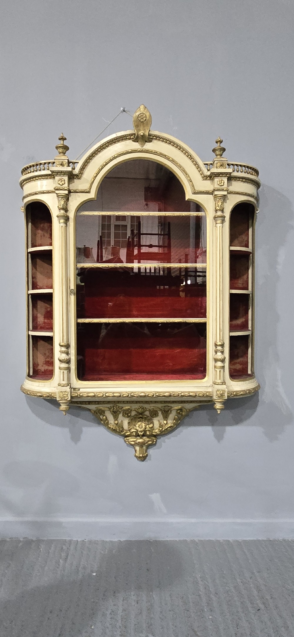 An antique French cream and gold painted wall vitrine featuring a central arched glazed door, open side shelves backed with red velvet, and carved gilt accents. View XIII