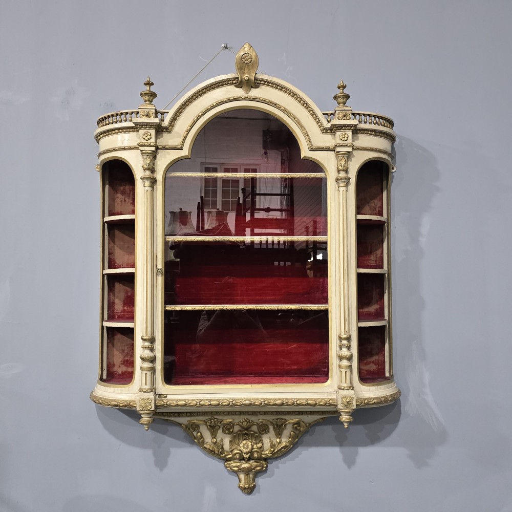 An antique French cream and gold painted wall vitrine featuring a central arched glazed door, open side shelves backed with red velvet, and carved gilt accents.
