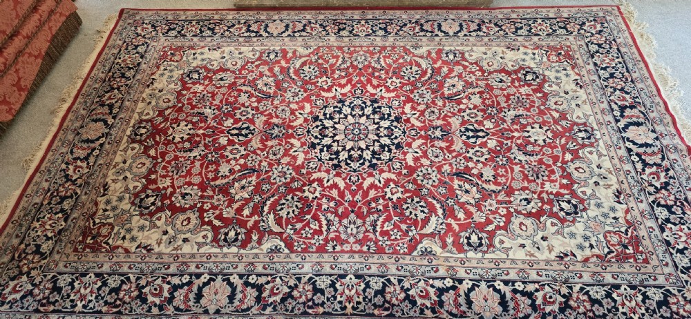 A large rectangular wool area rug featuring a central floral medallion on a red field surrounded by intricate navy and cream ornamental borders with fringed edges. View II