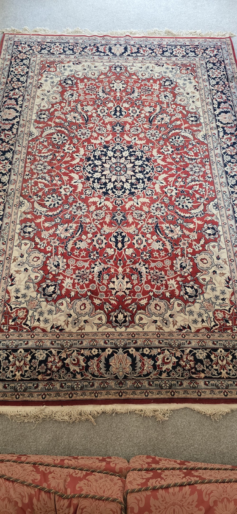 A large rectangular wool area rug featuring a central floral medallion on a red field surrounded by intricate navy and cream ornamental borders with fringed edges. View III