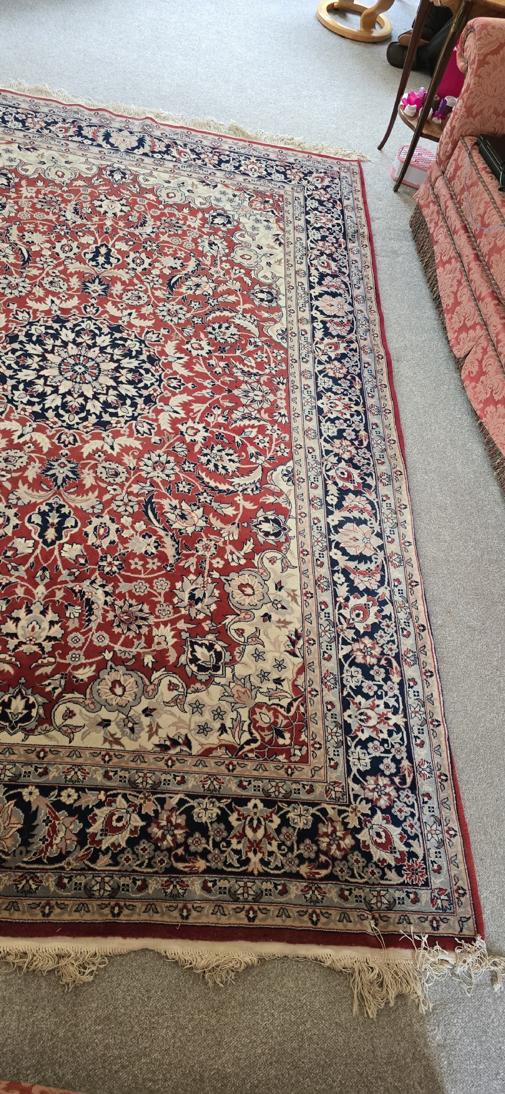 A large rectangular wool area rug featuring a central floral medallion on a red field surrounded by intricate navy and cream ornamental borders with fringed edges. View V