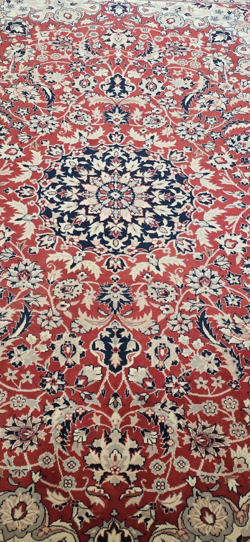 A large rectangular wool area rug featuring a central floral medallion on a red field surrounded by intricate navy and cream ornamental borders with fringed edges. View VII