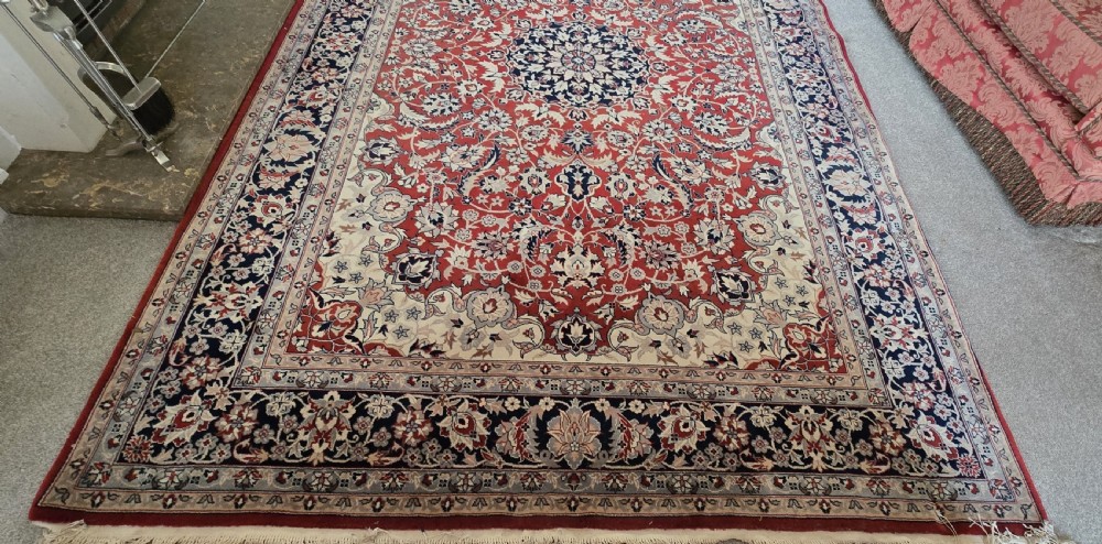 A large rectangular wool area rug featuring a central floral medallion on a red field surrounded by intricate navy and cream ornamental borders with fringed edges. View VIII