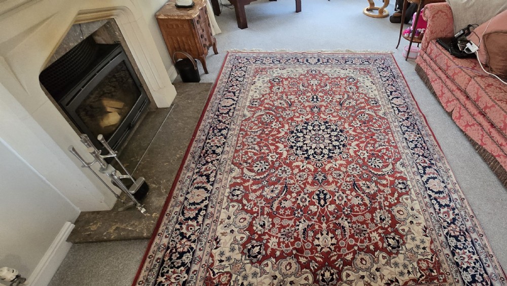 A large rectangular wool area rug featuring a central floral medallion on a red field surrounded by intricate navy and cream ornamental borders with fringed edges. View IX