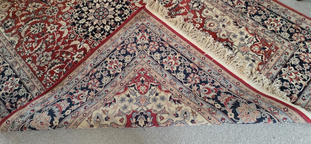 A large rectangular wool area rug featuring a central floral medallion on a red field surrounded by intricate navy and cream ornamental borders with fringed edges. View X