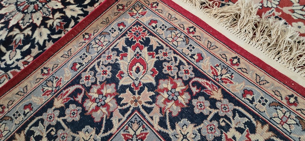 A large rectangular wool area rug featuring a central floral medallion on a red field surrounded by intricate navy and cream ornamental borders with fringed edges. View XI