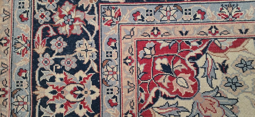 A large rectangular wool area rug featuring a central floral medallion on a red field surrounded by intricate navy and cream ornamental borders with fringed edges. View XII