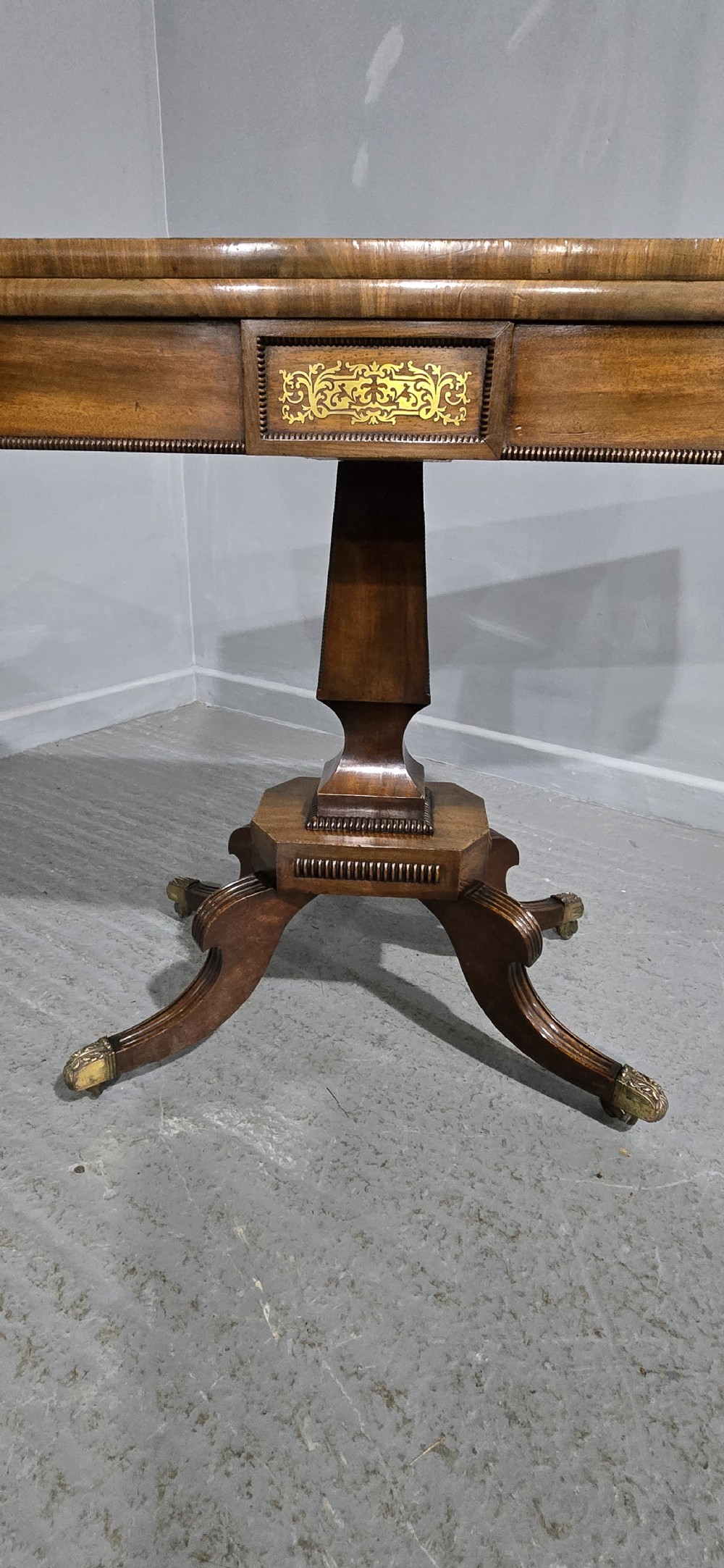 A Regency brass inlaid wooden card table featuring a fold-over top, central pedestal column, and four splayed legs with brass paw feet. View IX