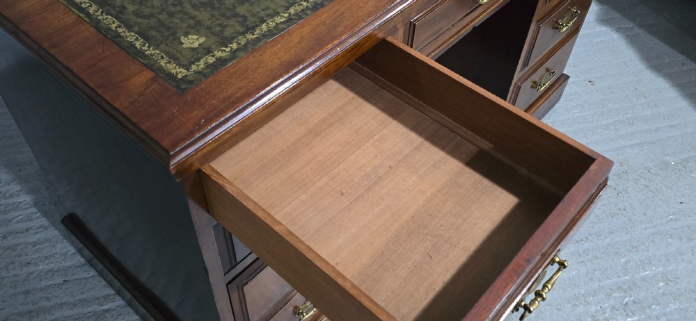 A warm brown mahogany pedestal desk featuring a green leather writing top, nine drawers, and brass swan-neck handles set upon twin plinth bases. View IX