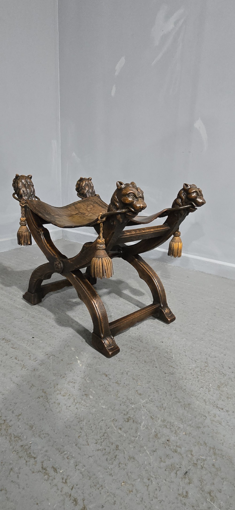 A carved dark walnut X-frame curule stool featuring lion heads with carved wooden tassels at the top corners and a slung brown leather seat. View V