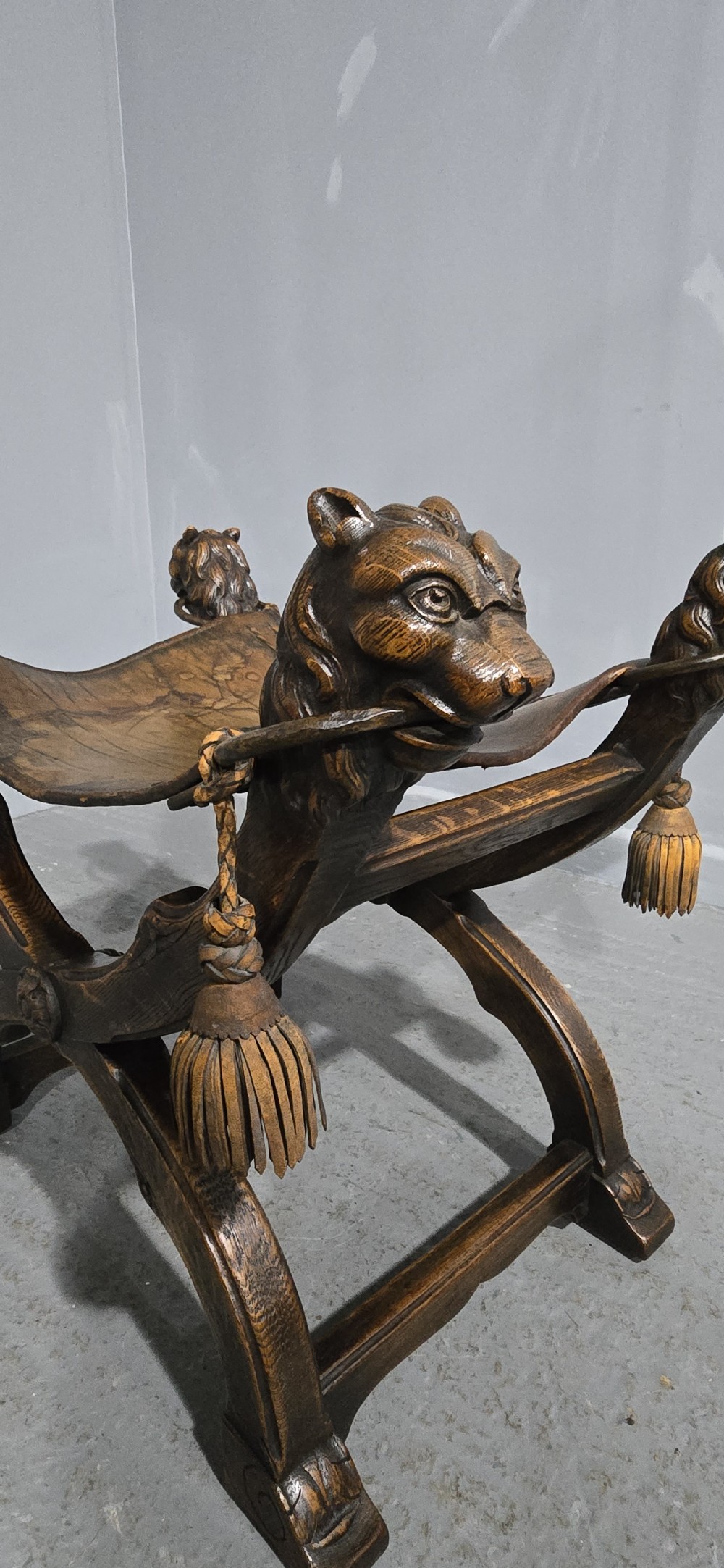 A carved dark walnut X-frame curule stool featuring lion heads with carved wooden tassels at the top corners and a slung brown leather seat. View VI