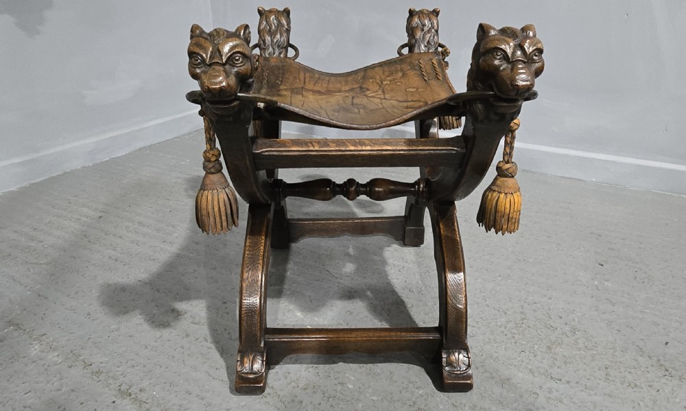 A carved dark walnut X-frame curule stool featuring lion heads with carved wooden tassels at the top corners and a slung brown leather seat. View VII